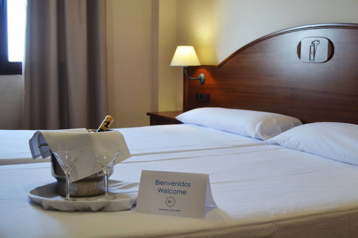 Hotellihuone, jossa on sänky, yöpöytälamppu, minibaari ja tervetulokyltti.