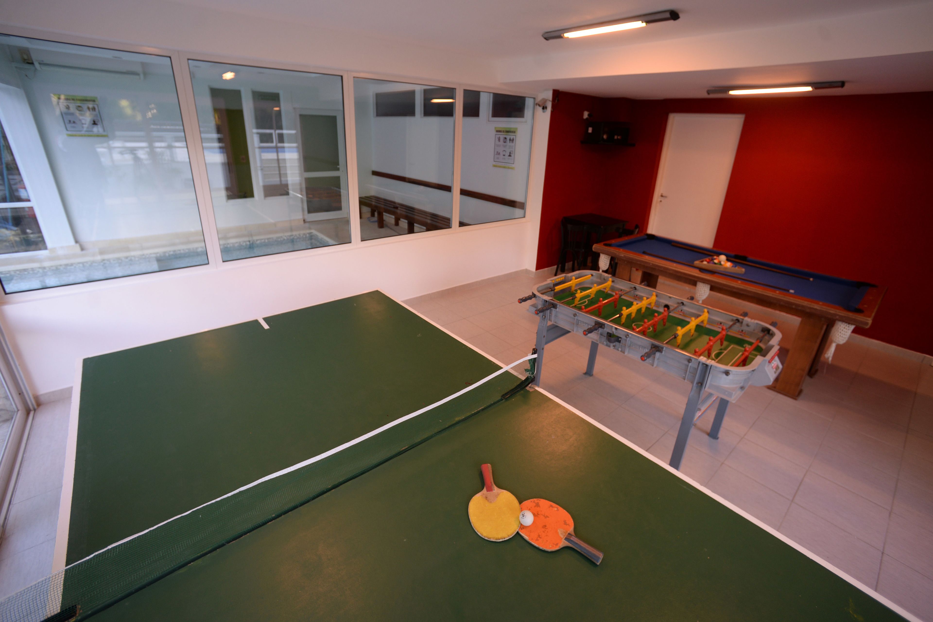 A ping pong table with two paddles and a ball, a foosball table, and a red wall.