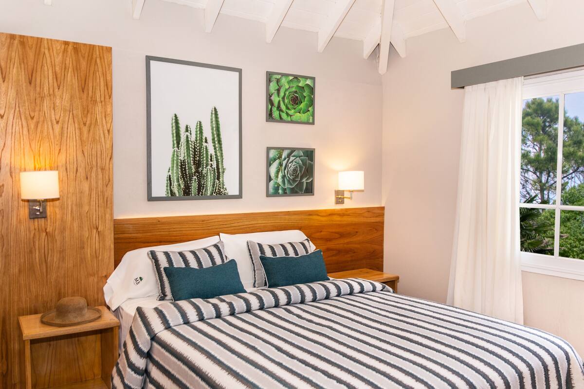 A neatly made bed with striped bedding, two wall-mounted lamps, and framed botanical artwork on the wall.