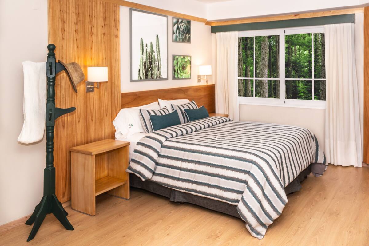 A bedroom with a wooden headboard, a bed with striped bedding, two bedside lamps, a wooden nightstand, and a window with a forest view.