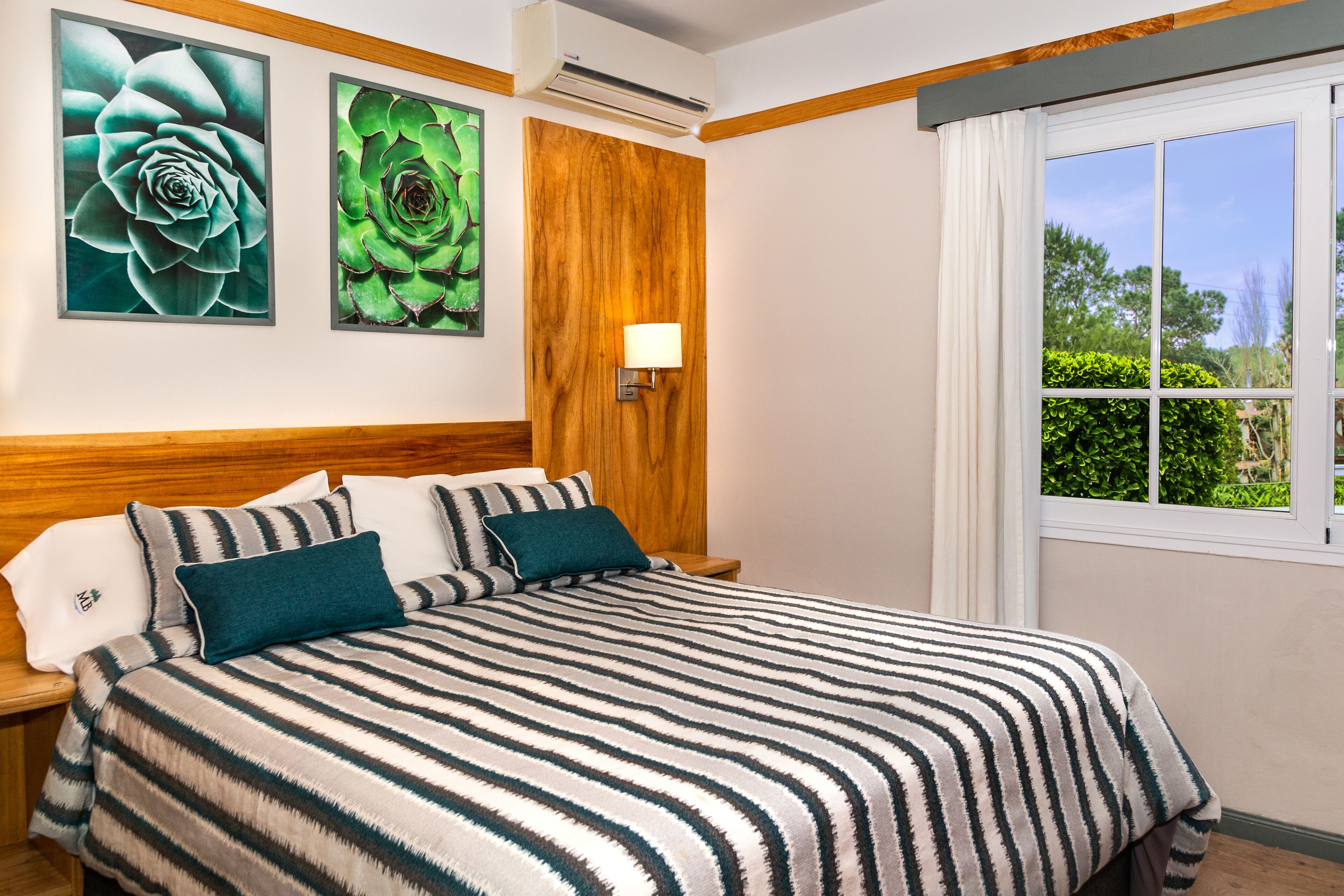 Hotelkamer met een groot bed, een houten hoofdbord, twee wandgemonteerde airconditioners, een raam met witte gordijnen, en twee ingelijste bloemkunstwerken aan de muur.