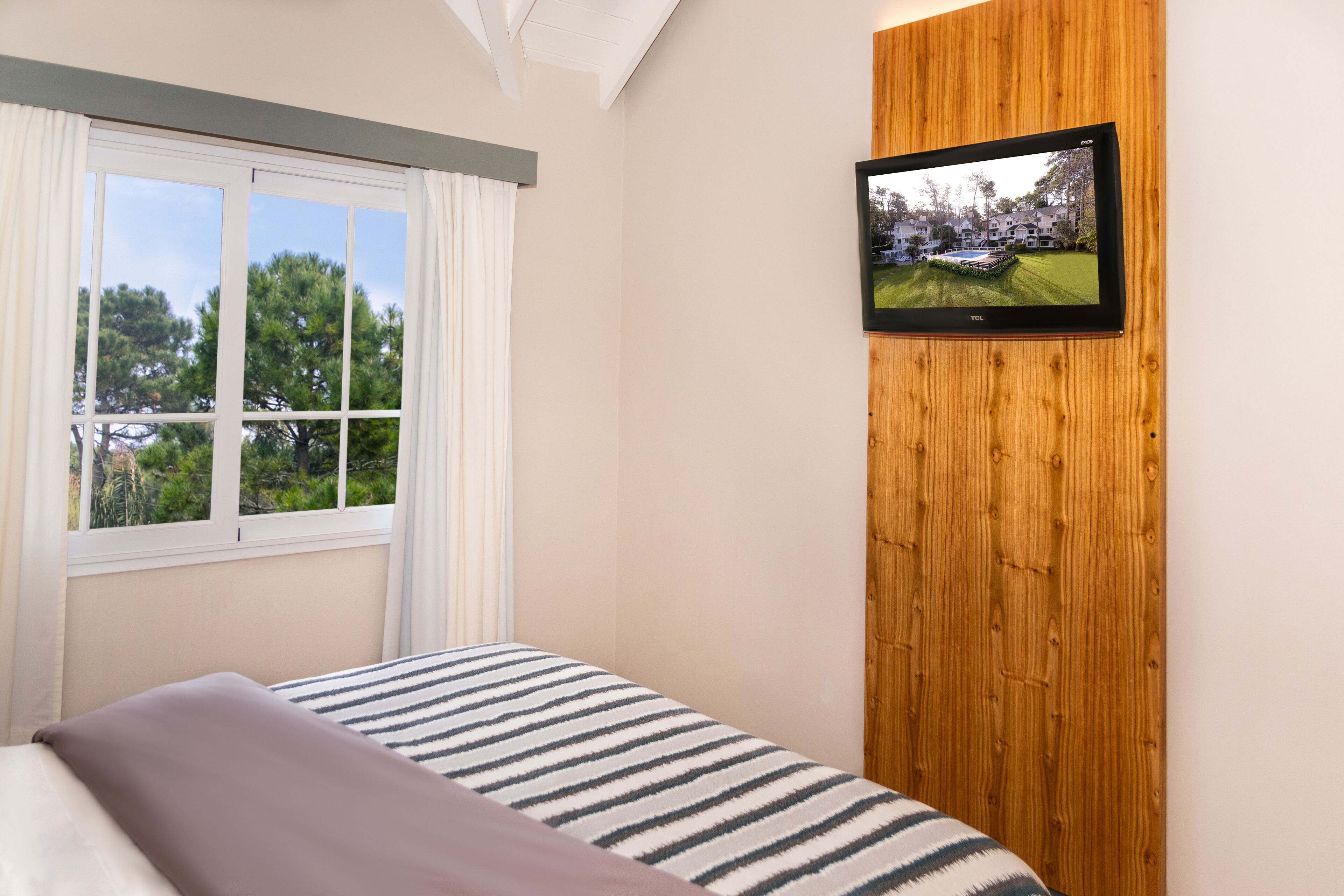 Quarto com cama, janela com cortinas, televisão fixada em um painel de madeira, e vista para árvores e edifícios lá fora.