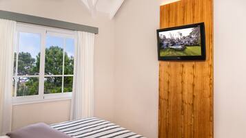 Quarto com cama, janela com cortinas, televisão fixada em um painel de madeira, e vista para árvores e edifícios lá fora.