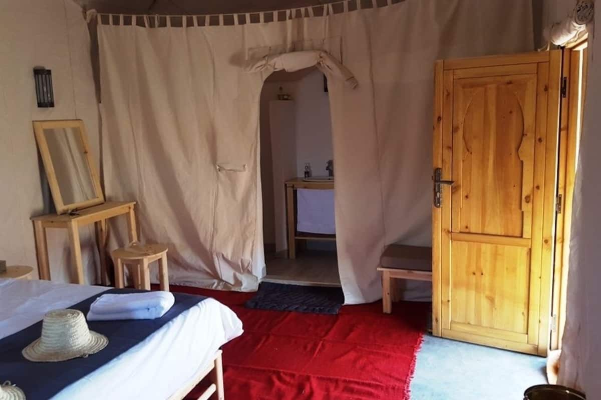 Quarto com cama, porta de madeira, espelho e uma mesinha.