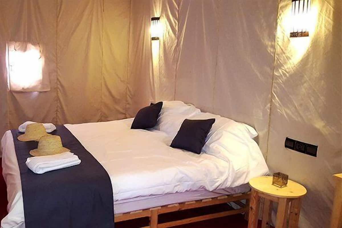 Uma cama de solteiro com lençóis brancos, duas almofadas pretas e um chapéu dobrado ao lado.