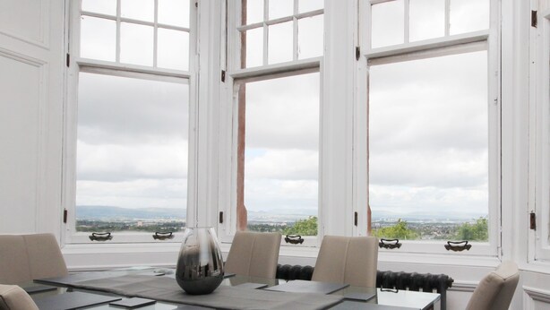 A dining area with a table set for six, large windows with a view of the city and mountains, and a modern vase centerpiece. - Braemount House (Paisley)