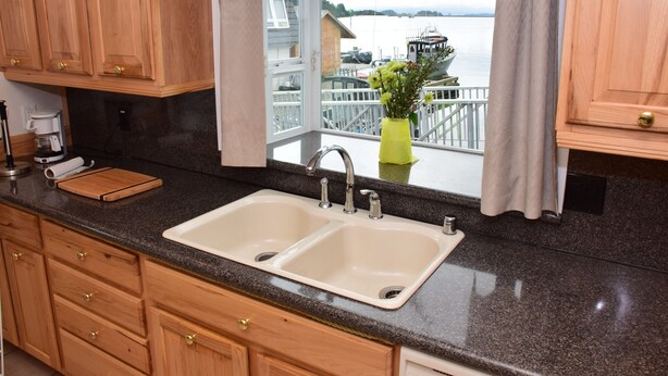 A kitchen with a double sink, wooden cabinets, and a view of a marina through a window. - Longliner Lodge and Suites (Sitka)