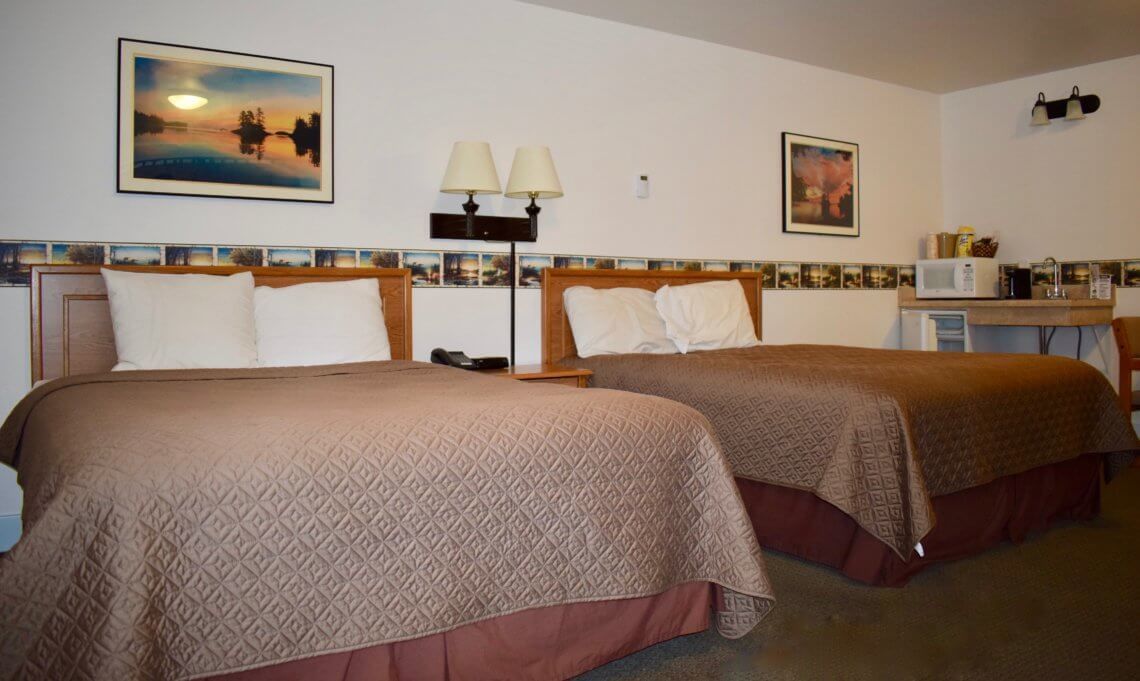 A hotel room with two beds, a small kitchenette, and a view of a lake at sunset.