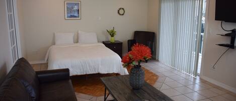 A bedroom with a bed, a wooden table with a vase and remote, a black chair, and a TV on the wall.