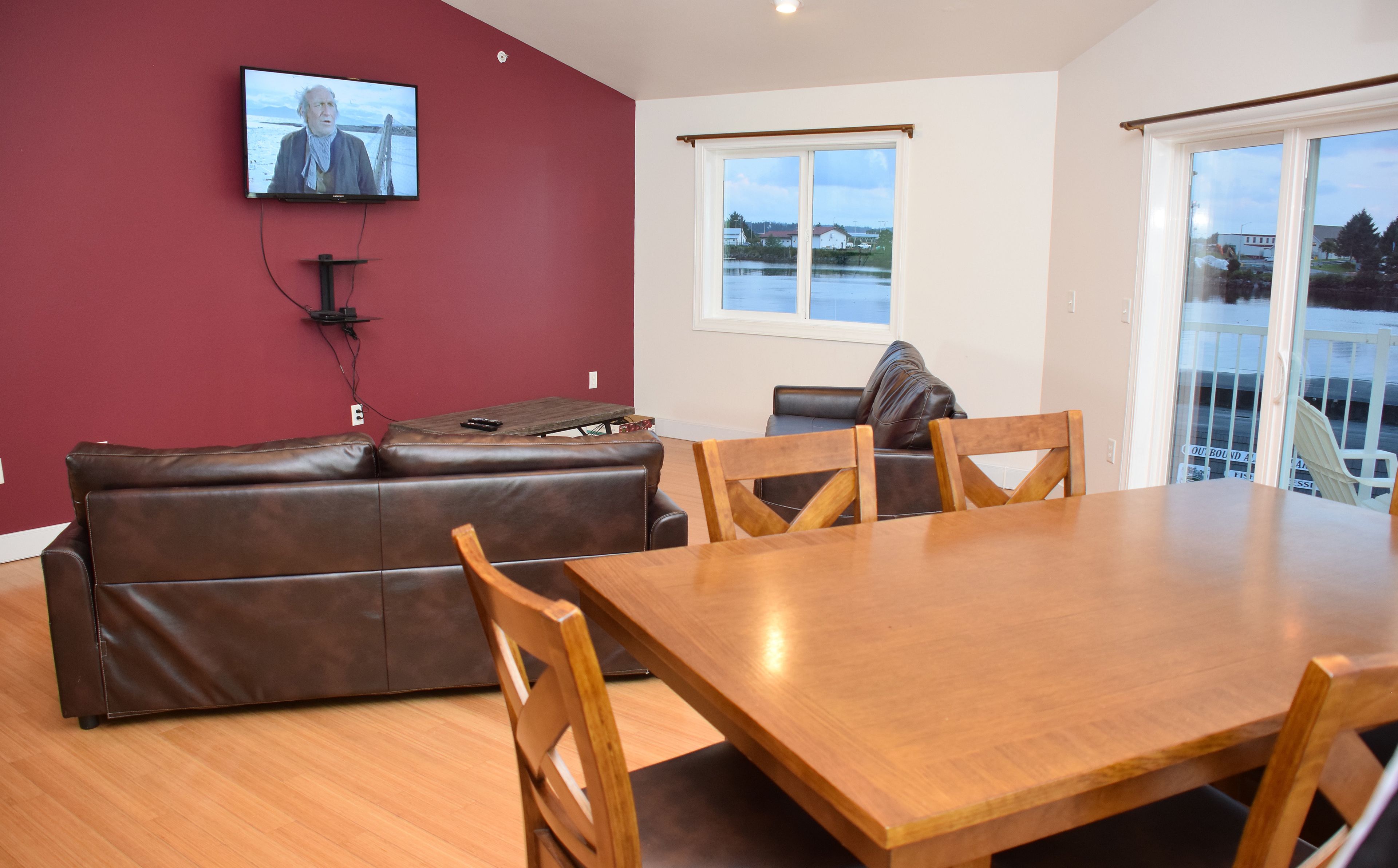 A living room with a brown leather sofa, a wooden dining table, and a television mounted on the wall.