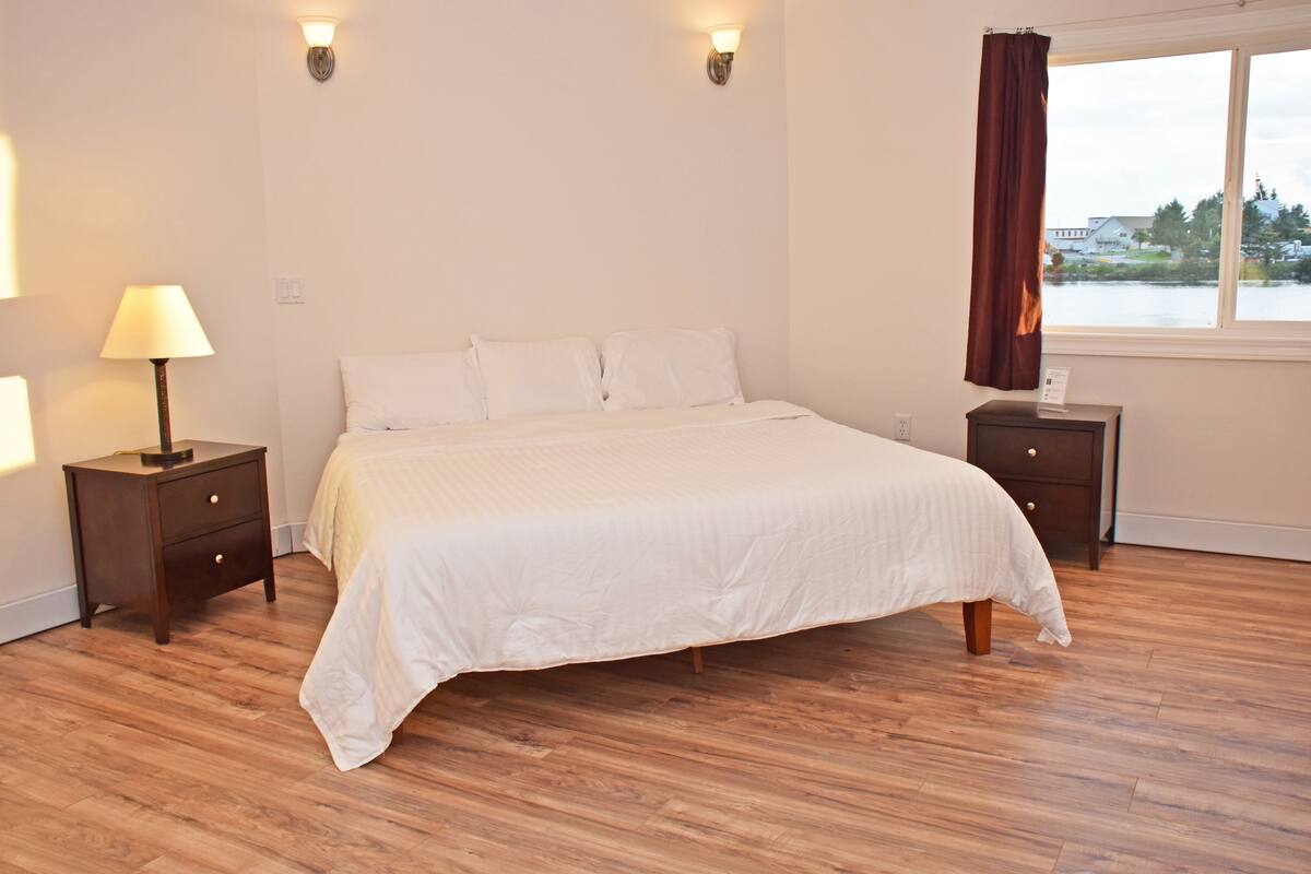 A bedroom with a large bed, two nightstands, a lamp, and a window with a view of a body of water.