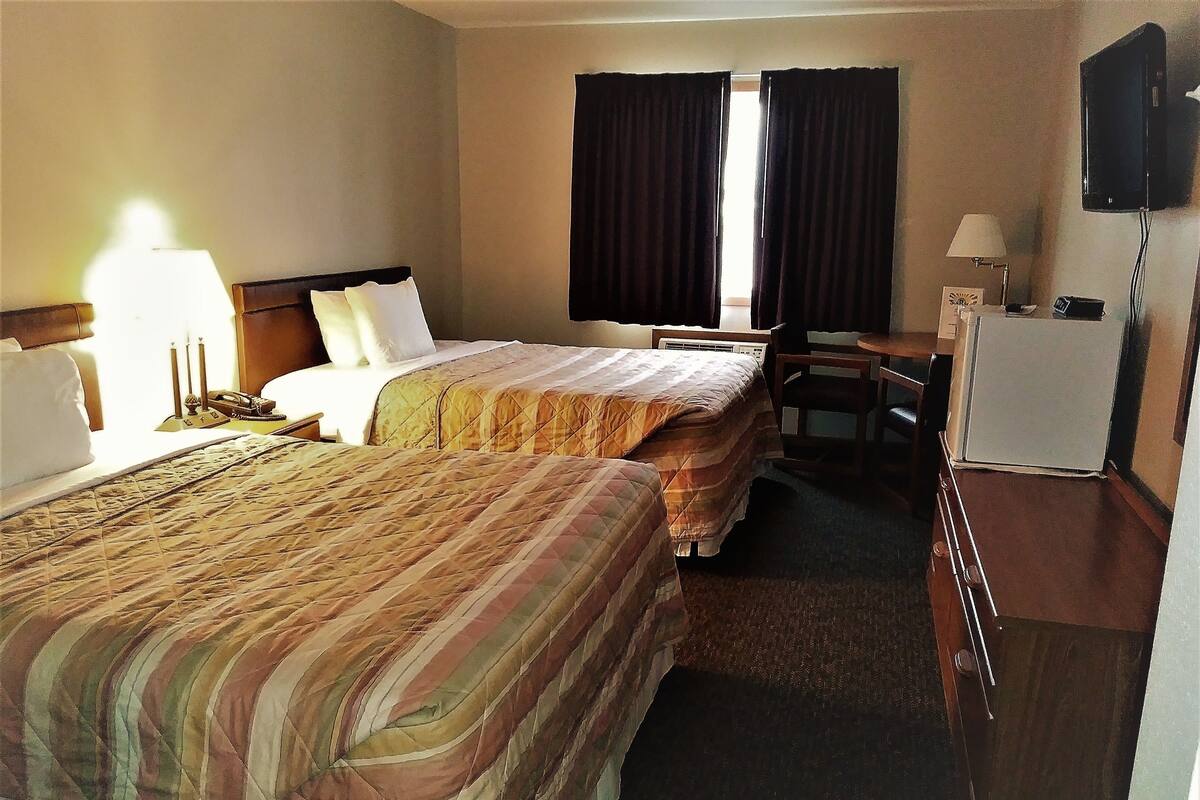 A hotel room with two beds, a desk, a chair, a refrigerator, and a television.