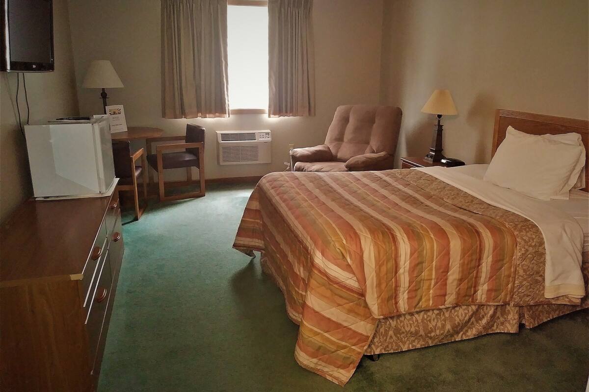A hotel room with a bed, a desk, a chair, a TV, and an air conditioning unit.