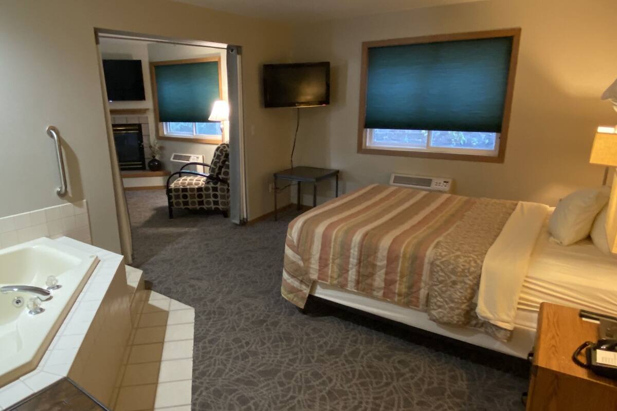 A hotel room with a large bed, a separate bathroom with a bathtub, a television mounted on the wall, and a small table with a lamp.