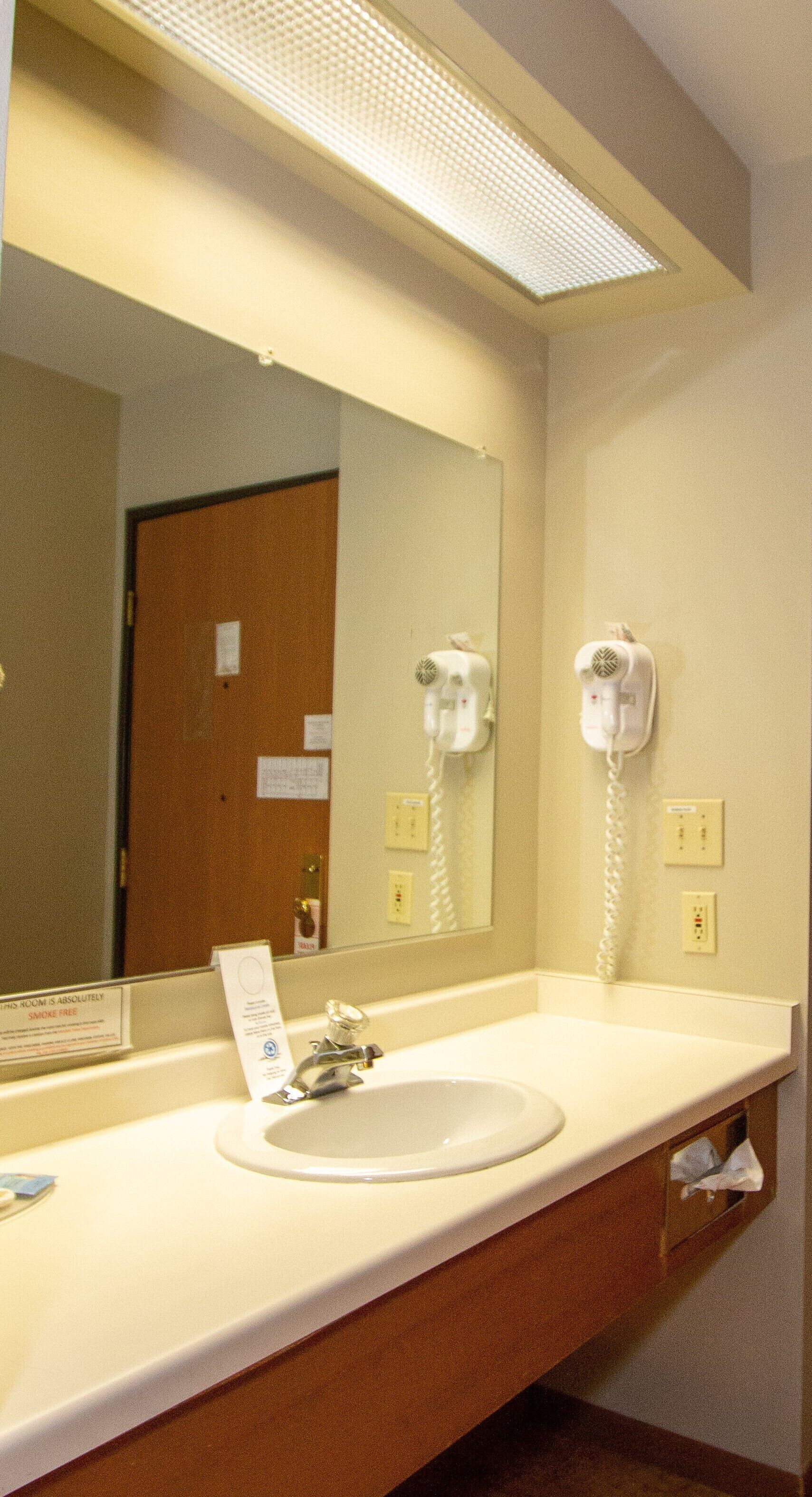 Une salle de bain d’hôtel équipée d’un lavabo, d’un miroir, d’un sèche-cheveux et d’un panneau indiquant que la chambre est non-fumeur.