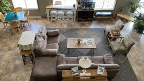 A spacious living room with a sectional sofa, a coffee table, and a piano. There is a dining area with a table and chairs, and a large window with blinds. - SunRise Inn Hotel (Mondovi)