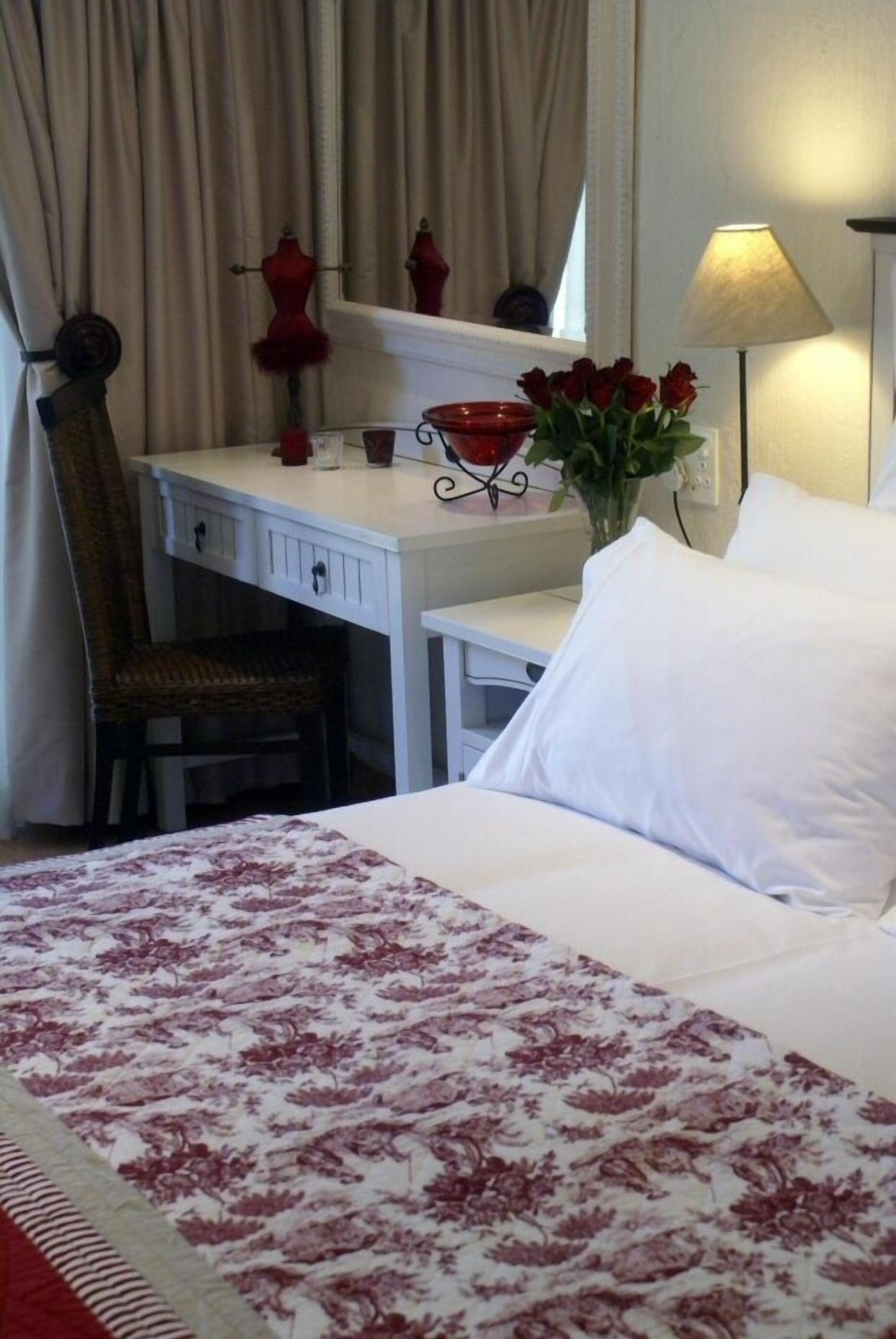Een hotelkamer met een bed, een bureau met stoel, een lamp, een vaas met rode rozen en een spiegel aan de muur.