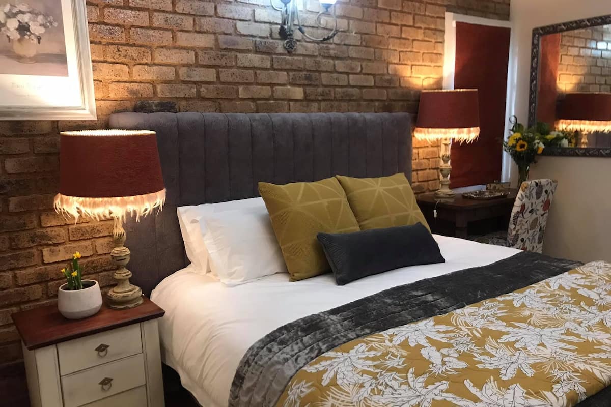 Ein Schlafzimmer mit Backsteinwand, einem Bett mit weißer und gemusterter Bettwäsche, zwei Nachttischlampen, einem Nachttisch und einem Spiegel.