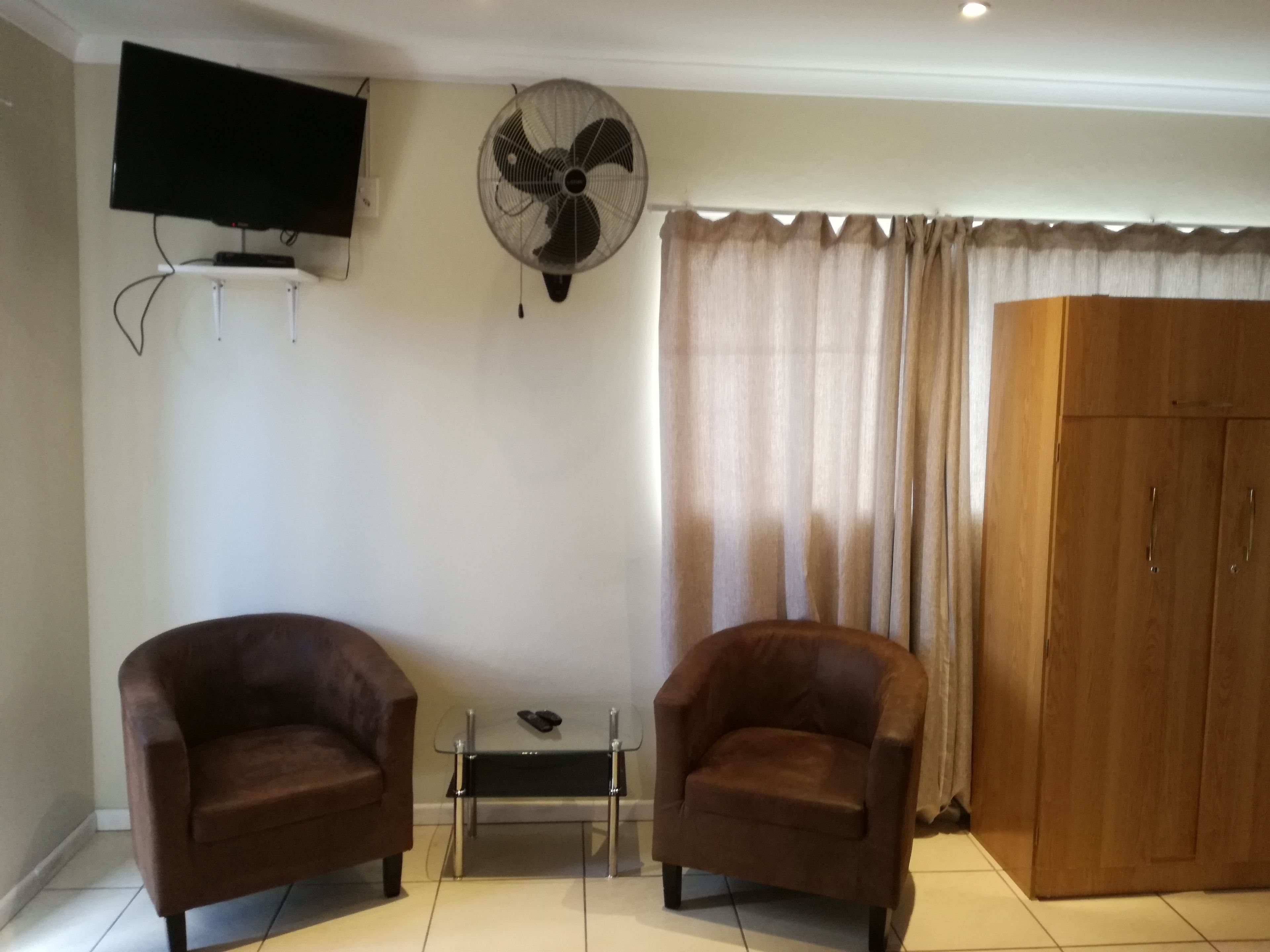 A room with two brown armchairs, a small table with a remote control, a wall-mounted fan, a television, and a large wooden wardrobe.