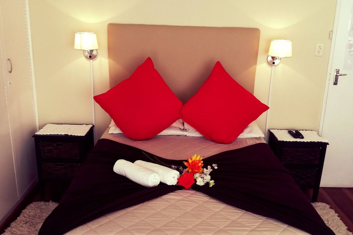 A hotel room with a bed, two red pillows, a white duvet, a black bed skirt, two white towels, a small flower arrangement, and two bedside lamps.