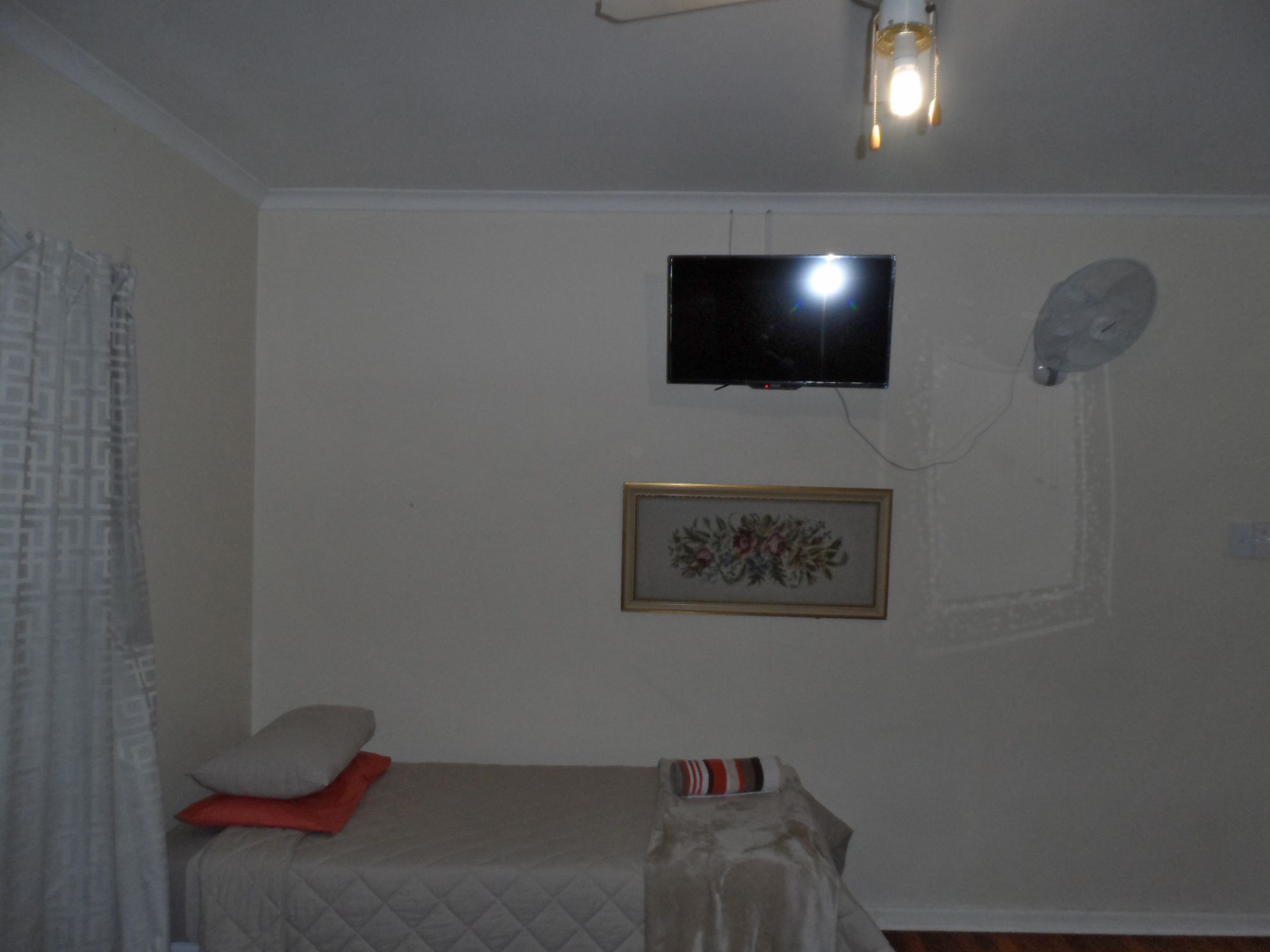 A small bedroom with a bed, a television mounted on the wall, a framed picture, and a ceiling fan.
