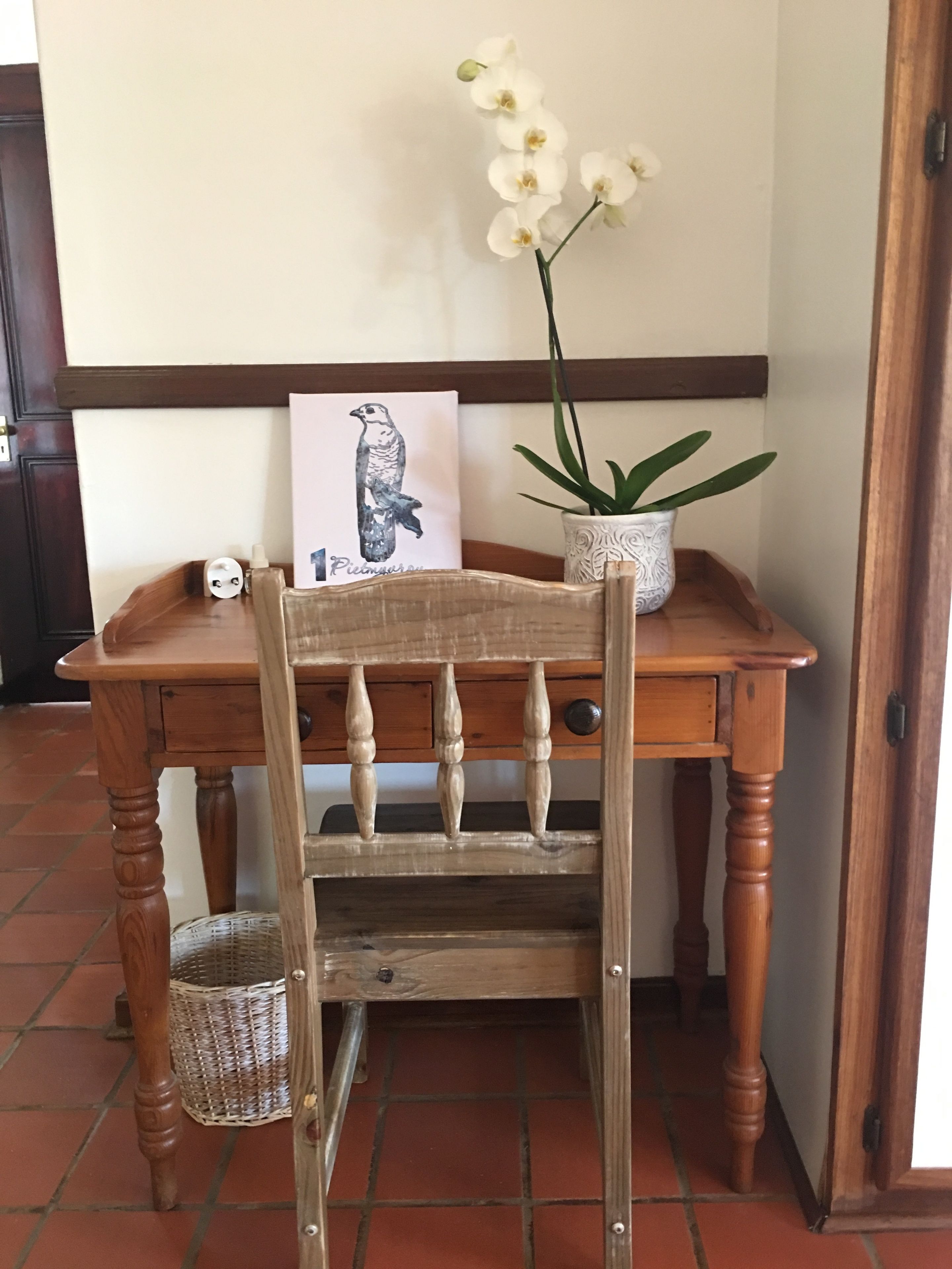 Una silla de madera con una cesta tejida debajo, una mesa con un cuadro enmarcado y una orquídea en maceta, y una puerta al fondo.