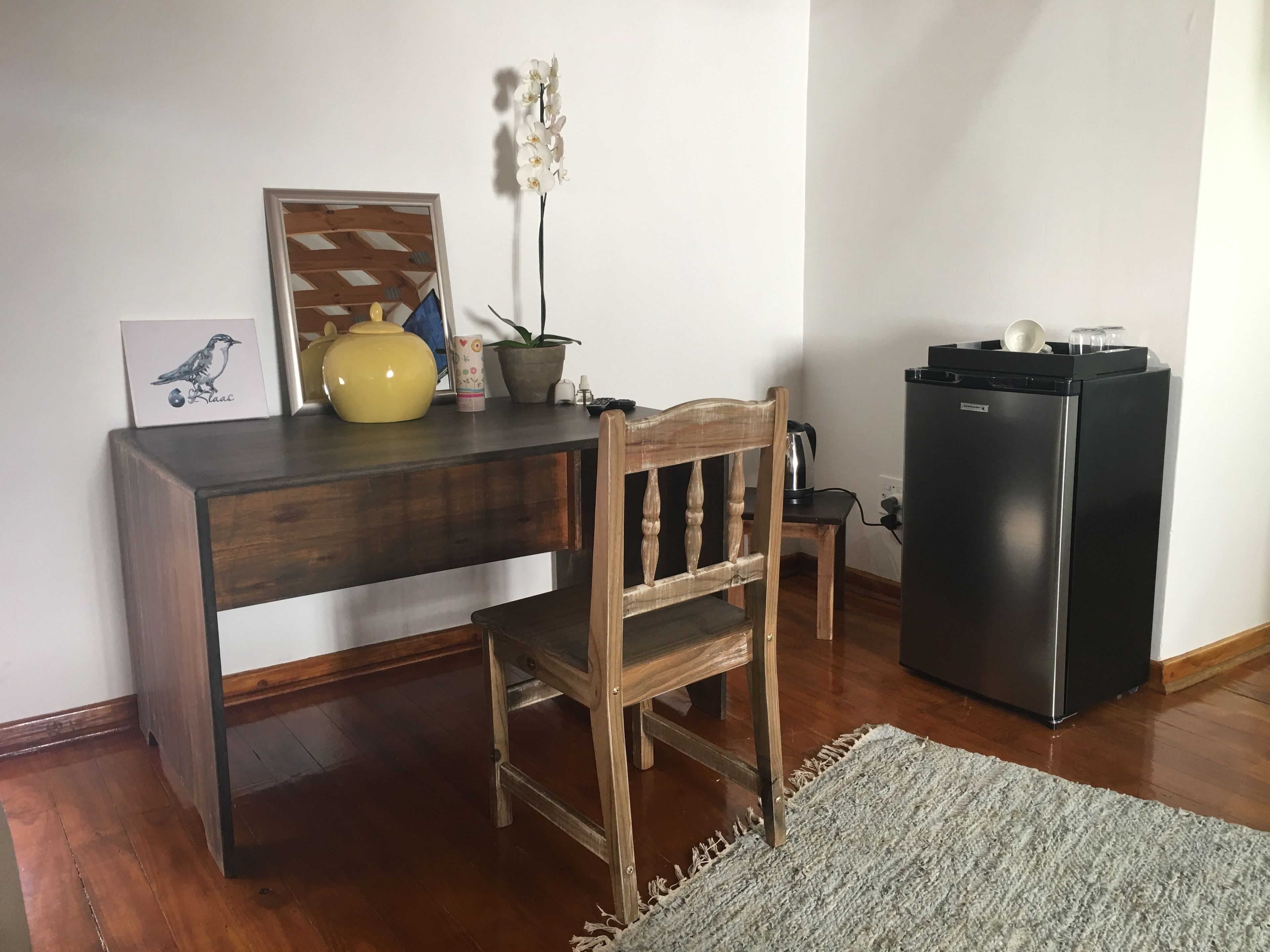 Un escritorio de madera con una silla, un refrigerador negro, una planta en maceta y un cuadro en la pared.