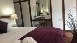 Una cama bien hecha con ropa de cama blanca y morada, una lámpara en la mesita de noche, una silla, un espejo y una planta en maceta.