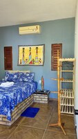 Quarto com cama, ar condicionado fixado na parede, um quadro colorido e uma estante de madeira.