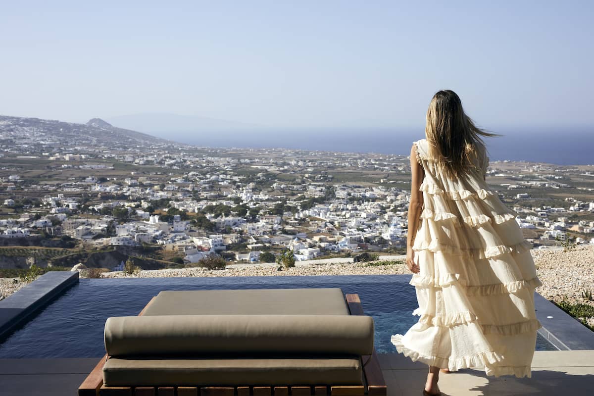 A woman in a white dress standing by a pool overlooking a coastal town.