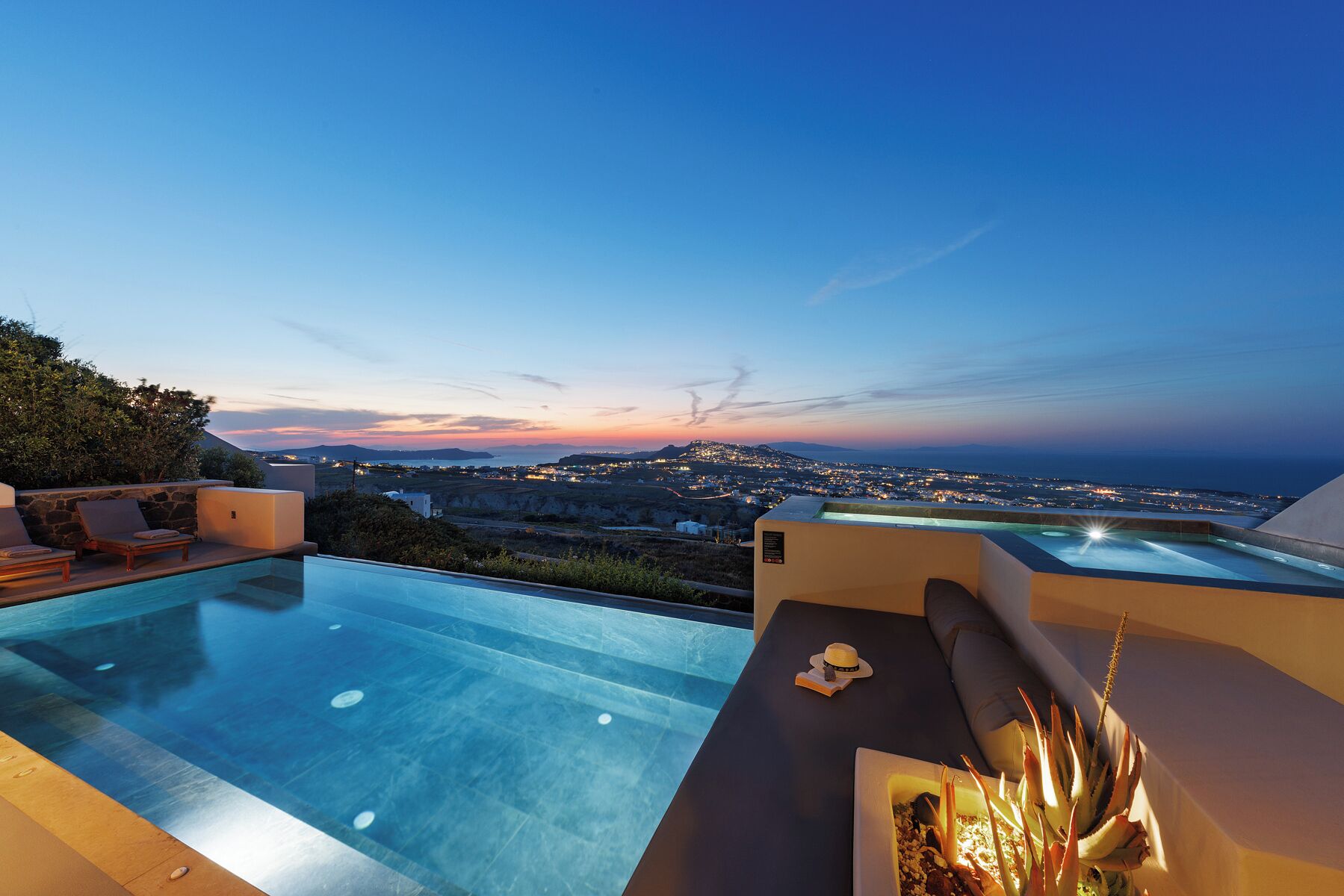 Un espace aménagé au bord de la piscine, offrant une vue sur la ville au coucher du soleil, avec un fauteuil de relaxation et une table agrémentée d’une plante.