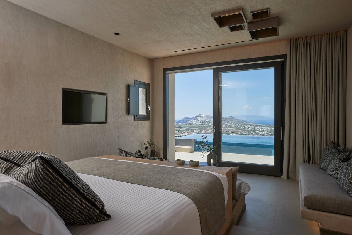 A modern hotel room with a large bed, a sofa, a TV mounted on the wall, and a sliding glass door leading to a balcony with a scenic view.
