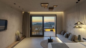 A modern hotel room with a large bed, a flat-screen TV, a dining area with a table and chairs, and a balcony with a view of the landscape. - North Santorini - A Luxury Spa Hotel (Santorini)