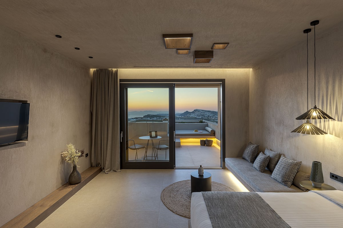 A modern hotel room with a large bed, a flat-screen TV, a dining area with a table and chairs, and a balcony with a view of the landscape.