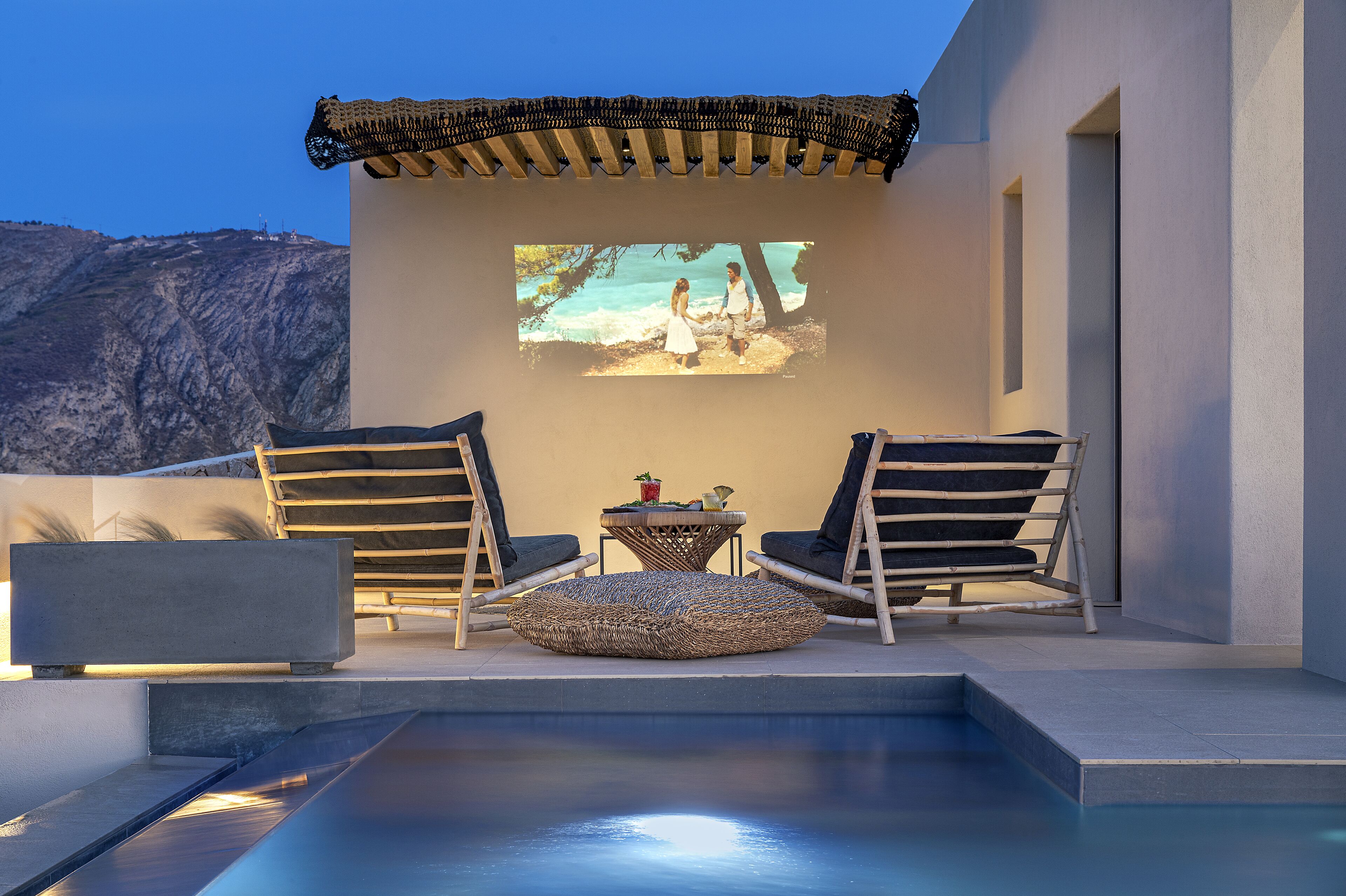 Un espacio de estar moderno al aire libre con dos sillas y una pequeña mesa, con vista a una piscina y un paisaje montañoso.
