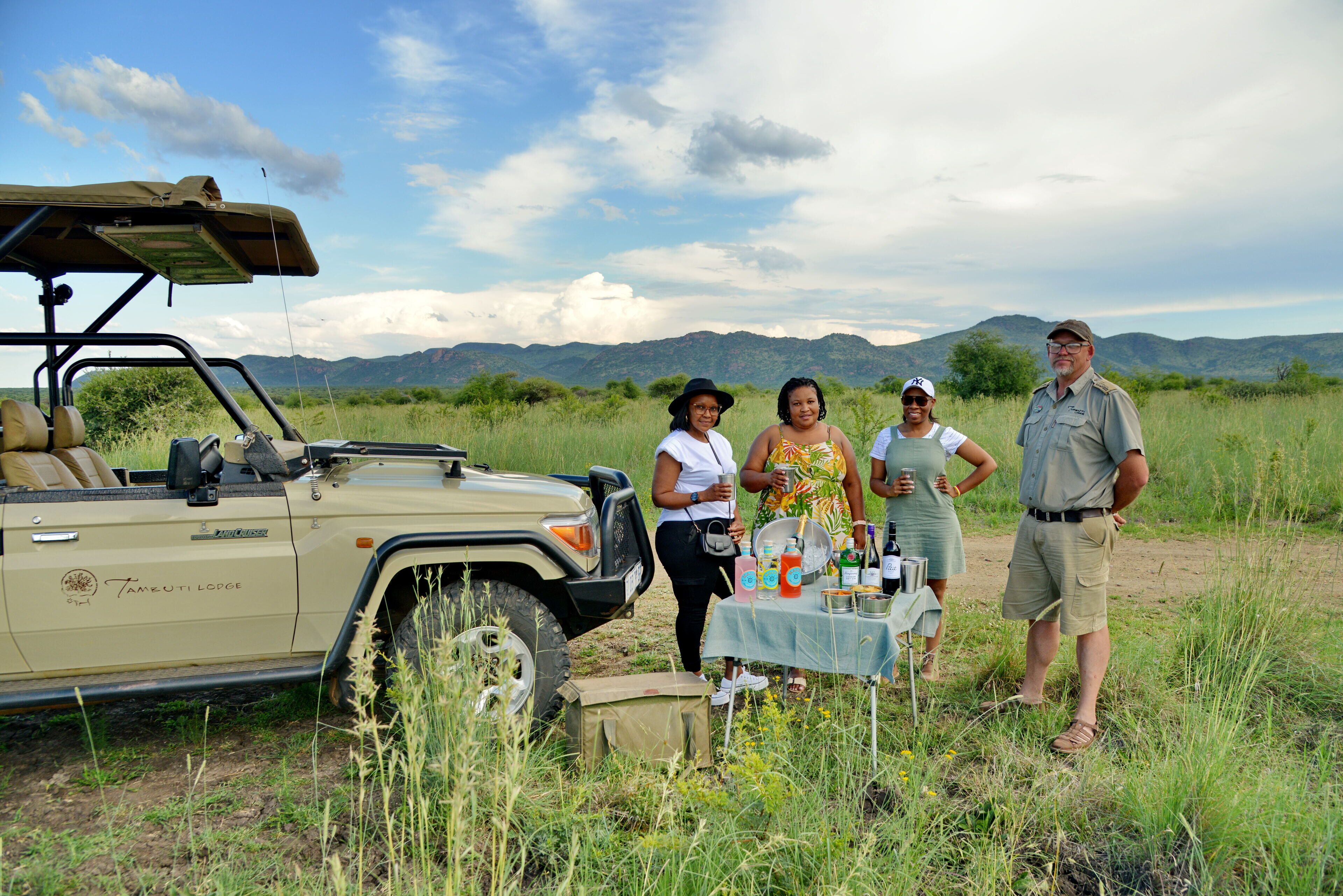 A safari vehicle with a table set up in a grassy field with mountains in the background.