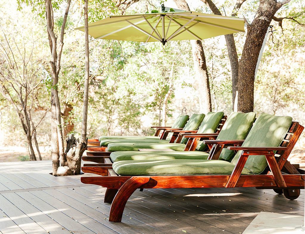 Une rangée de fauteuils de jardin en bois, dotés de coussins verts, sur une terrasse en bois, le tout entouré d’arbres.