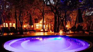 Ein kreisförmiger Pool mit einer zentralen Fontäne, umgeben von Liegestühlen und Sonnenschirmen, vor einer Kulisse aus Bäumen und einem Gebäude mit warmer Beleuchtung.