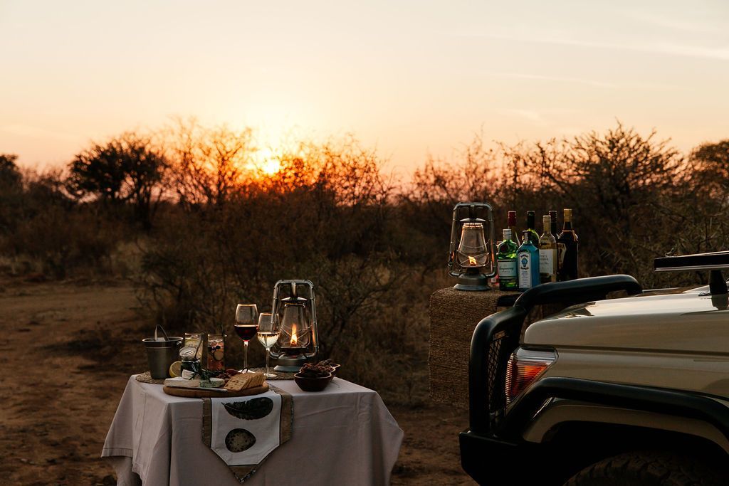 Mesa al aire libre con comida, bebidas y un farol, con el atardecer de fondo.