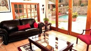 A living room with a black leather sofa, a glass coffee table, and a red armchair. A wooden door leads to a pool area. - Mizizi House of Sandton Bed & Breakfast (Sandton)