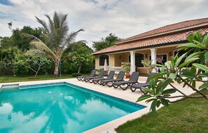 A swimming pool with lounge chairs, a covered patio, and lush greenery.