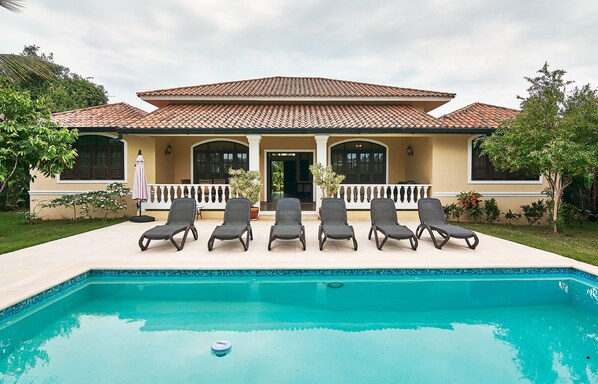 A house with a swimming pool, five lounge chairs, and a tiled roof.