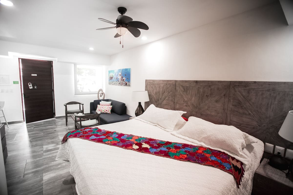 A modern hotel room with a large bed, a wooden headboard, a ceiling fan, a desk with a chair, and a window with a view.