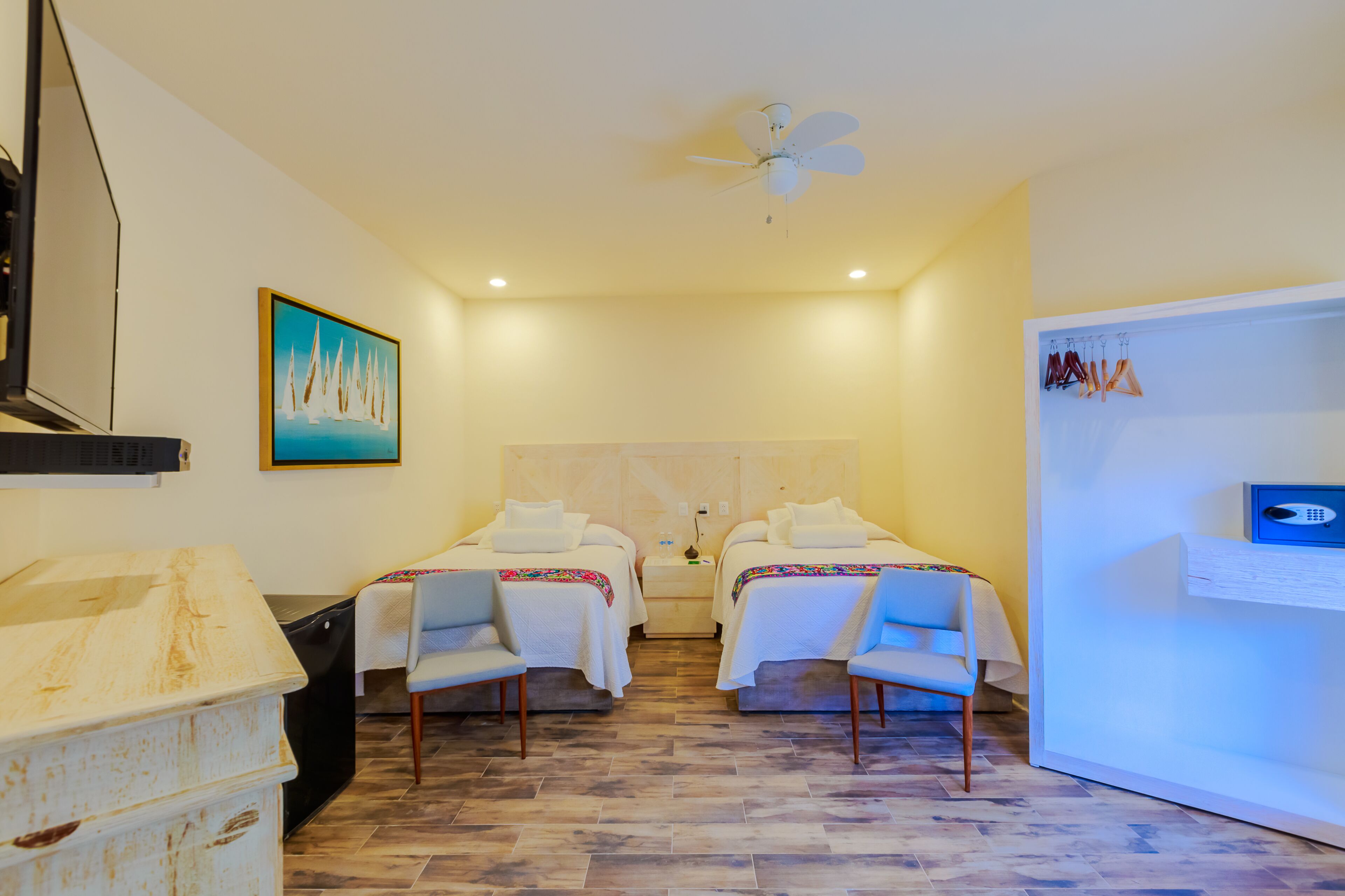 A hotel room with two beds, a ceiling fan, a painting, a desk with a computer, and a minibar.