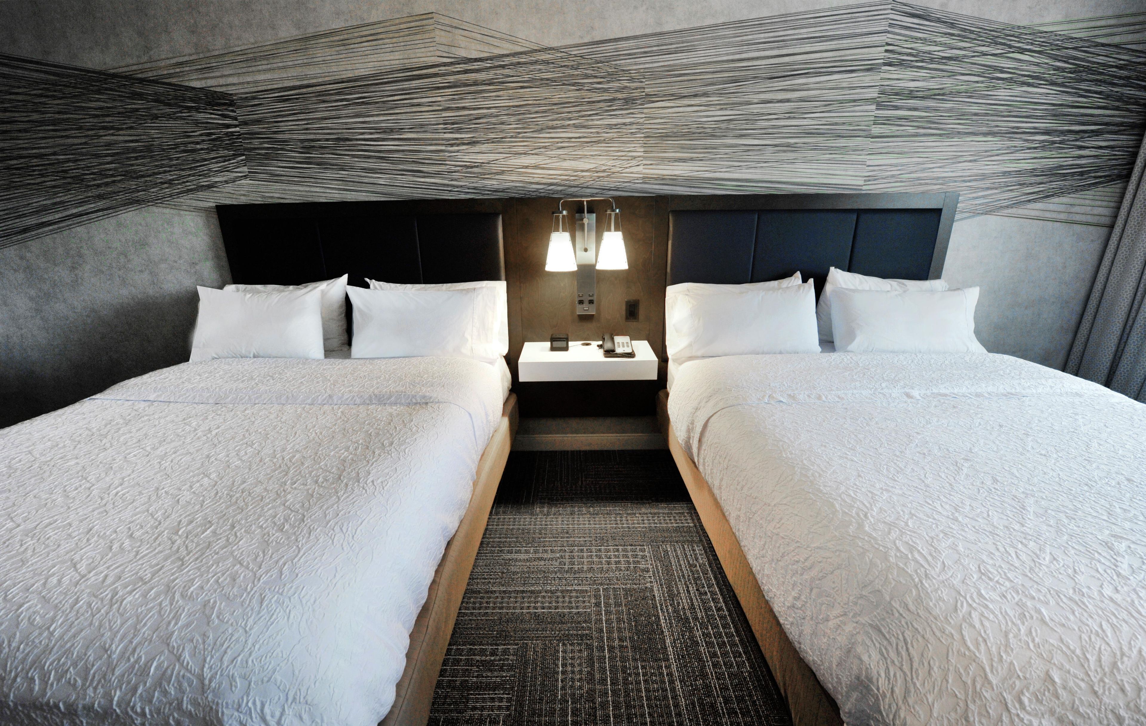 A hotel room with two beds, a dark headboard, a small table with a lamp, and a patterned carpet.