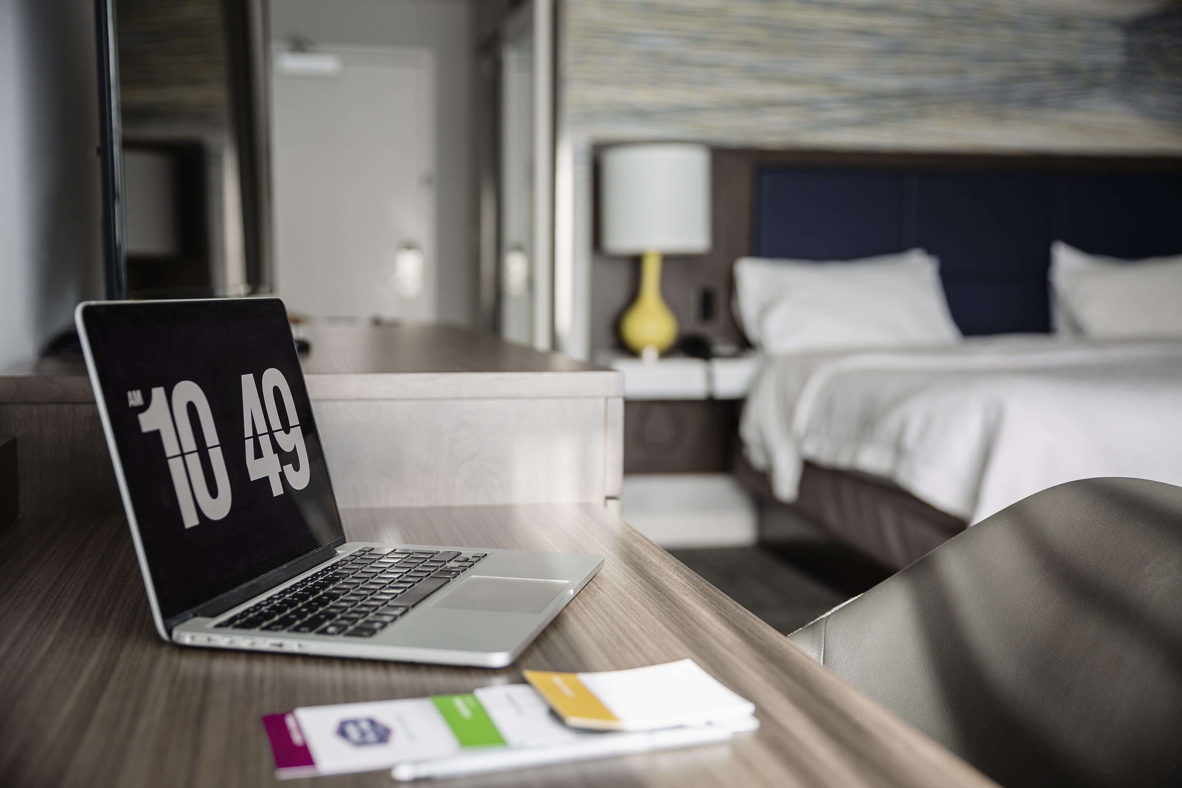 Ein Laptop auf einem Holztisch, im Hintergrund ein Hotelzimmer mit Bett und Nachttisch mit Lampe.