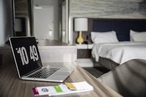 A laptop on a wooden desk with a hotel room in the background, including a bed and a nightstand with a lamp. - Hampton Inn & Suites by Hilton Quebec City Levis (Levis)