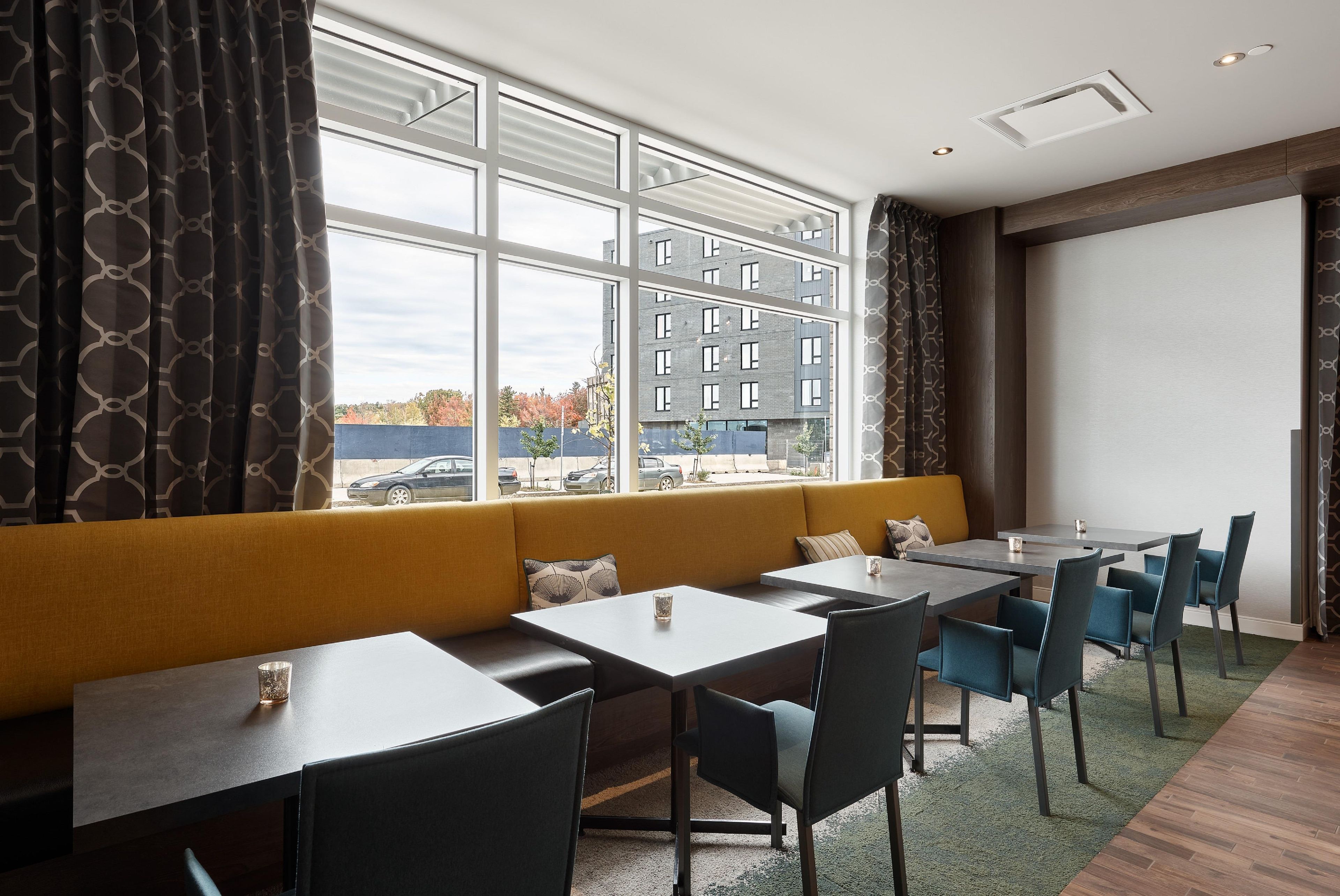 Una habitación de hotel moderna con un ventanal grande, un sofá y una zona de comedor con mesas y sillas.