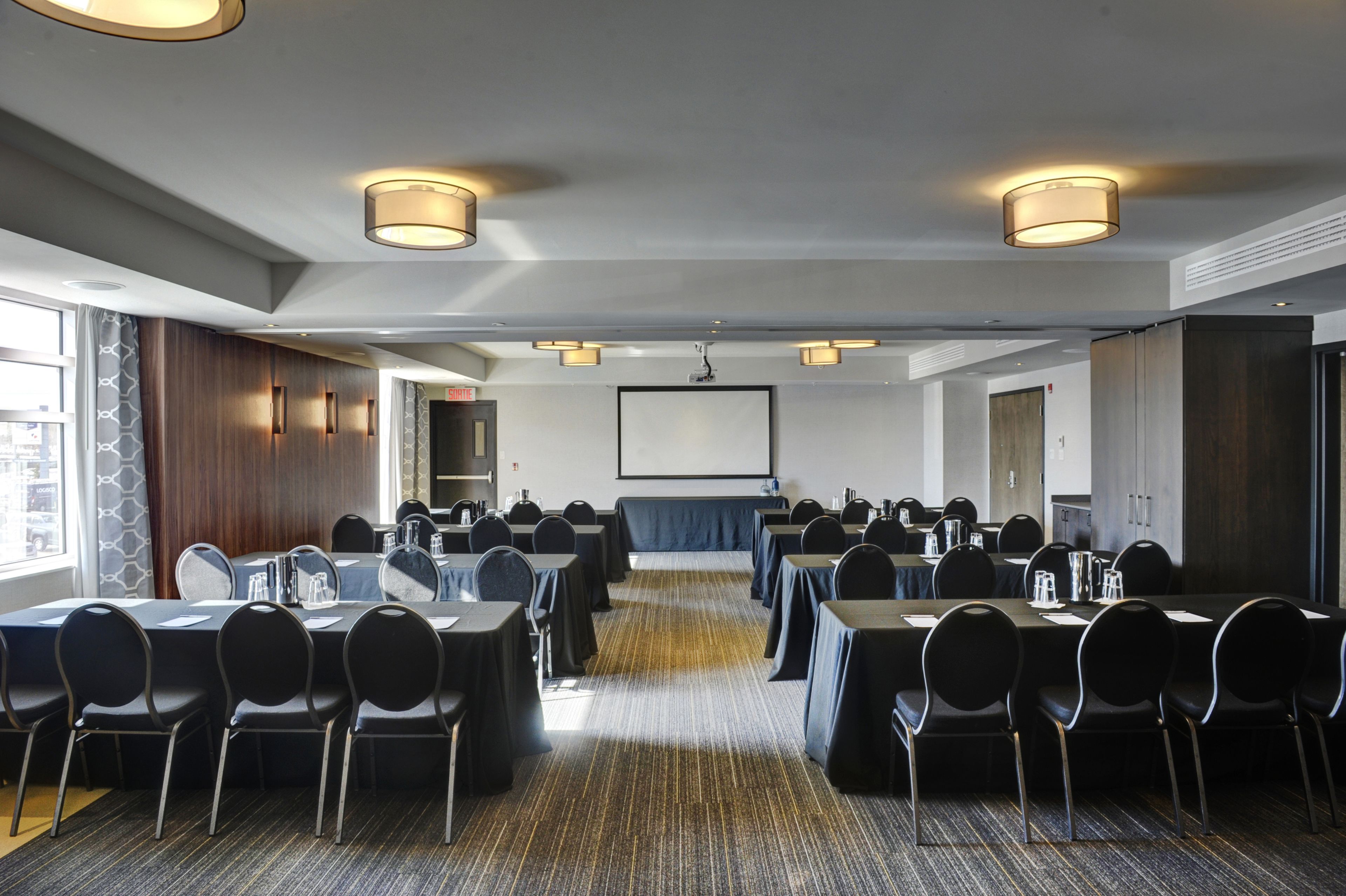 A conference room with multiple tables and chairs, a projector screen, and ceiling lights.