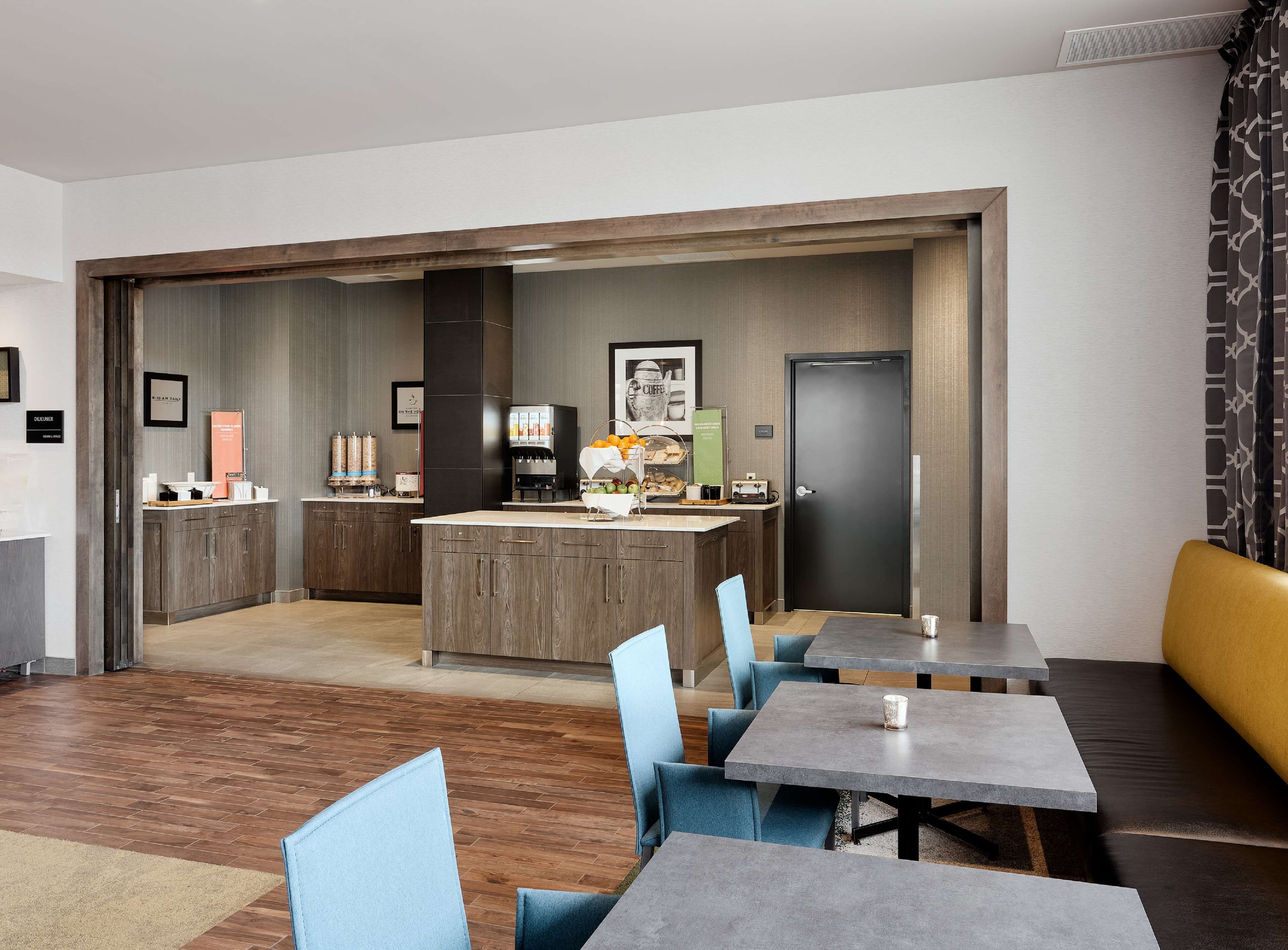 A modern hotel room with a kitchenette, a dining area with tables and chairs, and a separate entrance door.